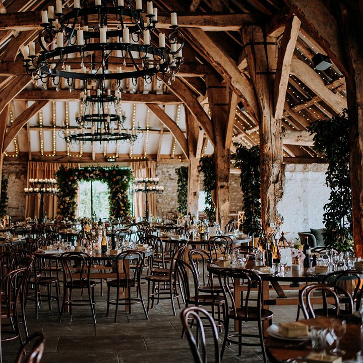 Rustic barn wedding reception complete with floral archway and column flowers, festoon lighting across wooden beams, and circular tables
