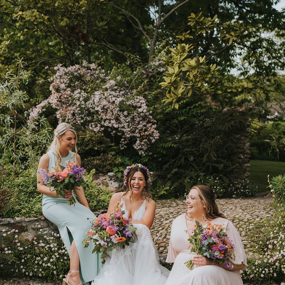 Bride sits with her bridesmaids in mismatched dresses holding colourful floral bouquets 