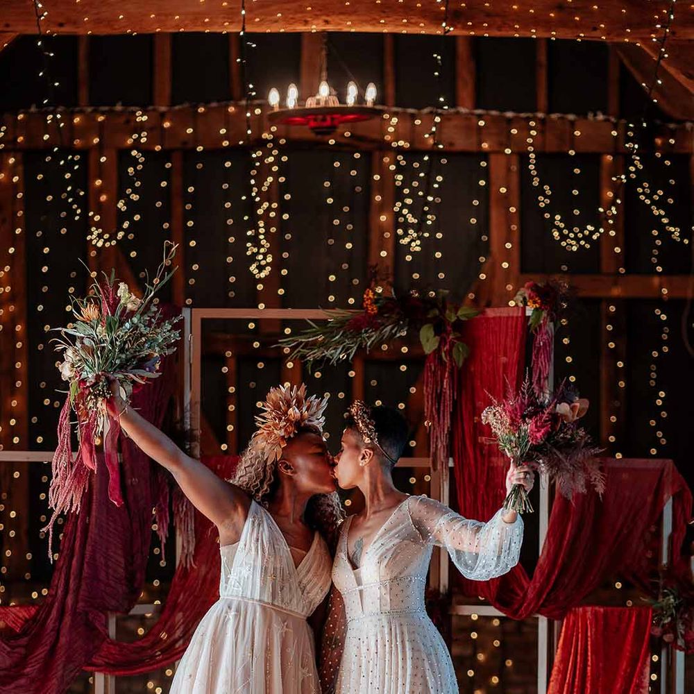 Bride in sleeveless floaty peach gown and gold leaf crown kissing bride in crystal embellished gown and gold headdress with red wedding drapes, foliage and dried flower decor and fairy lights in the background