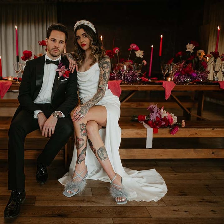 Groom in classic black tuxedo with magenta amaryllis and orchid boutonniere and bride in cowl neck sleeveless satin wedding dress with leg slit, gold glittery wedding headband, and soft grey fluffy feather open toe strappy high heels sitting on steps at Elmore Court with hot pink tapered candles, dark brass candlesticks, red roses, hot pink peonies and wine glasses 