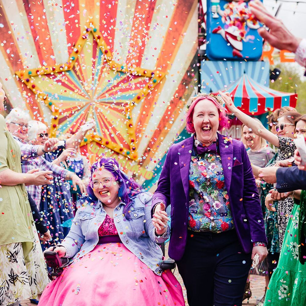 Bride in purple velvet blazer and black suit trousers with purple bowtie, pink patterned pocket square and colourful patterned shirt holding hands with bride in wheelchair wearing personalised denim jacket, pink wedding dress and blue tights doing colourful confetti throw 