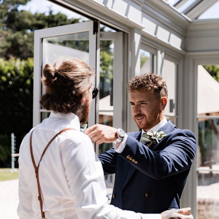 Groom in double breasted blazer helps groomsmen get ready on the morning of his wedding day