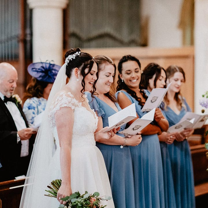 The bride stands with her bridesmaids in blue bridesmaid dresses reading out the hymns for the church wedding ceremony 