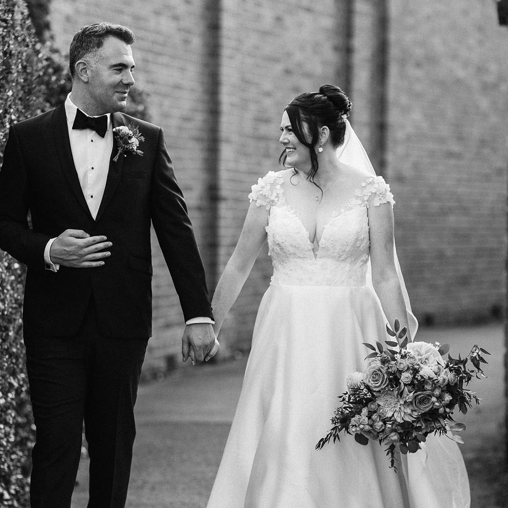 The bride wears a flower wedding dress with her brown hair in an elegant updo walking with the groom in a black tuxedo 
