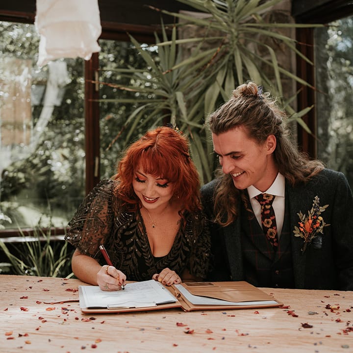 Groom in dark grey three piece suit and burgundy flower patterned tie and bride in black dress for wedding signing  
