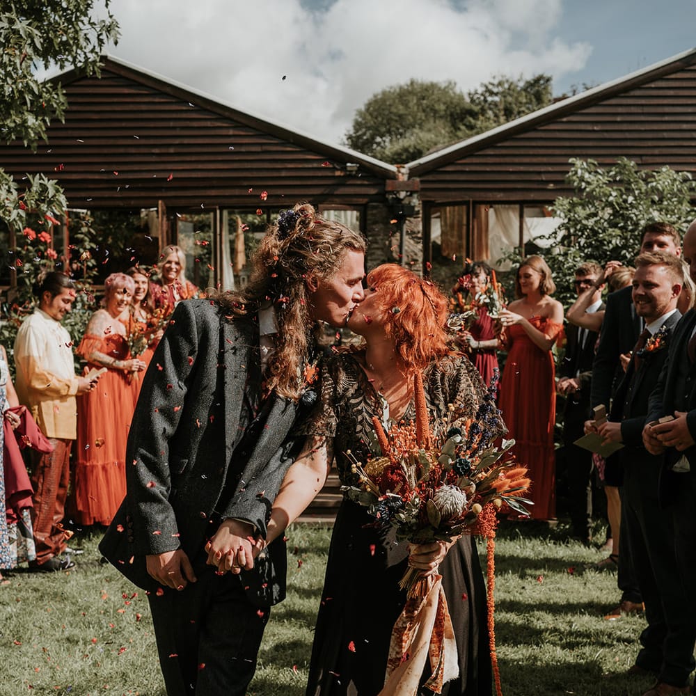 Groom in dark grey three piece suit and burgundy flower patterned tie and bride in embellished black wedding dress holding large orange dried flower bouquet kissing during wedding confetti exit 