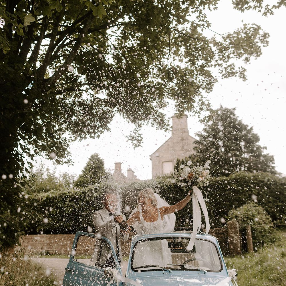 The bride and groom in a light blue vintage Fiat 500 wedding car decorated with ribbon and signage popping champagne to celebrate 