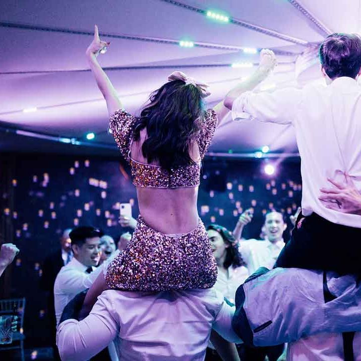 Bride in sparkly two piece bridal set and groom in white shirt and black trousers dancing on guests' shoulders 