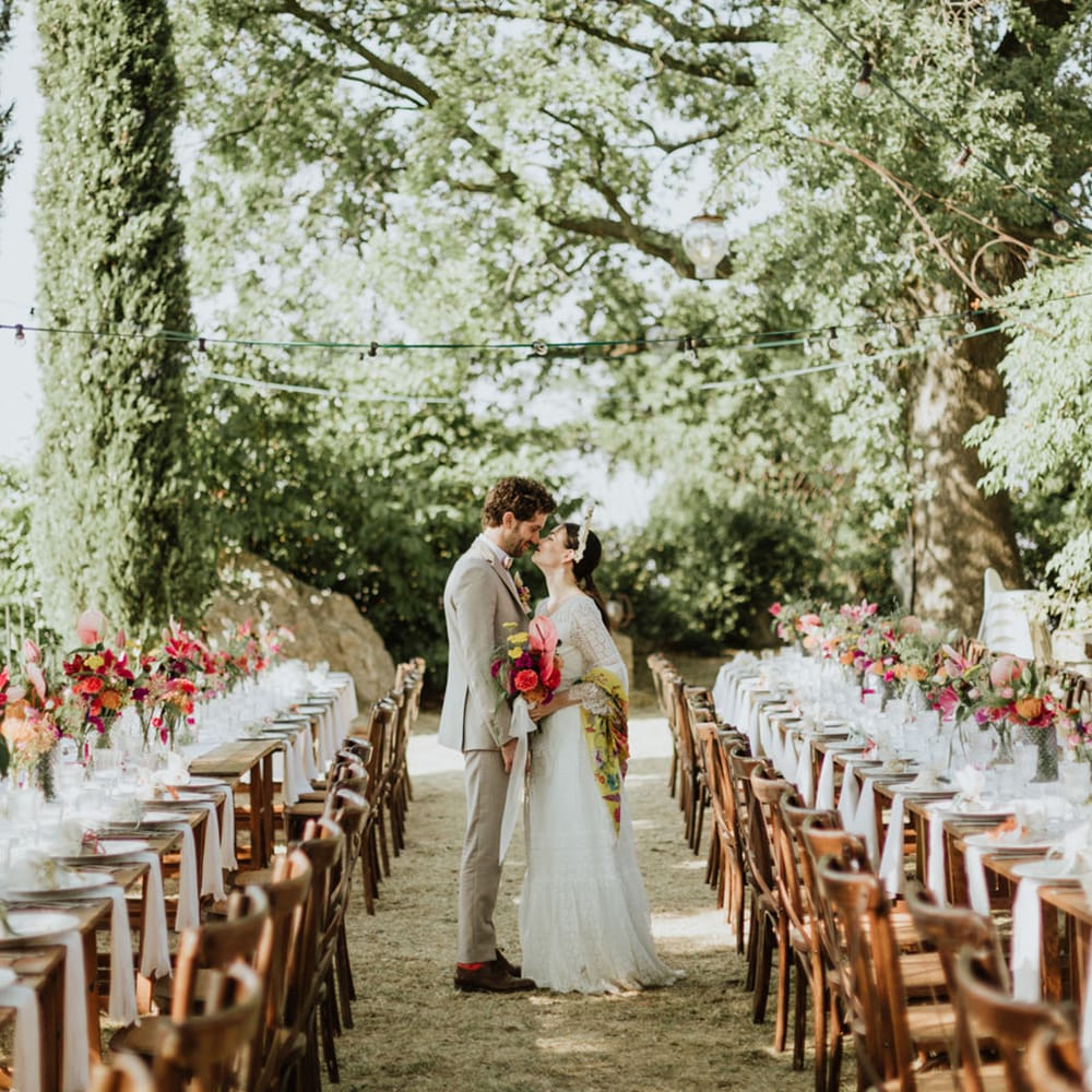 Bride in lace wedding dress and bridal crown with a pink bridal bouquet kissed groom in greige suit surrounded by wedding tables covered in pink floral decor