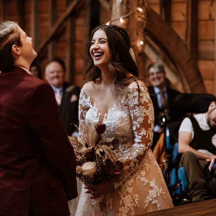 Bride in lace illusion sleeve wedding dress with overlay holding large autumnal boho dried flower bouquet laughing at the alter with bride in maroon bridal suit 