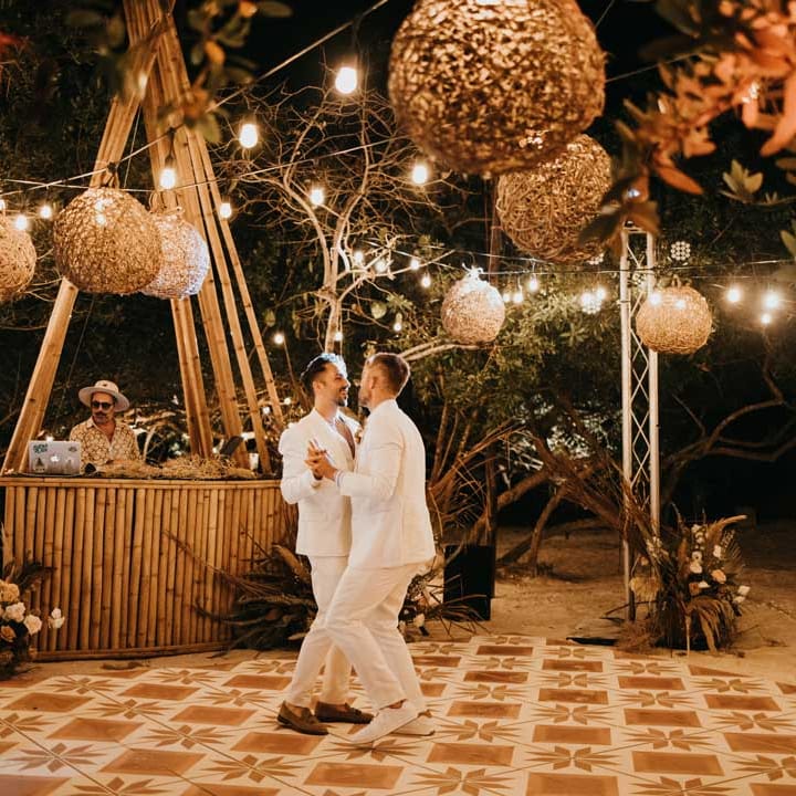 Grooms in matching white grooms suits doing their first dance as a couple on DIY hand painted wedding dance floor under twine lampshades and fairy lights 