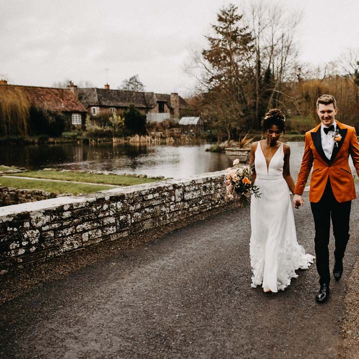 Bride in v neck sleeveless wedding dress with 3D floral applique train walking on the grounds of Brinsop Court wedding venue with groom wearing crushed burnt orange velvet grooms blazer, black bowtie and black suit trousers 