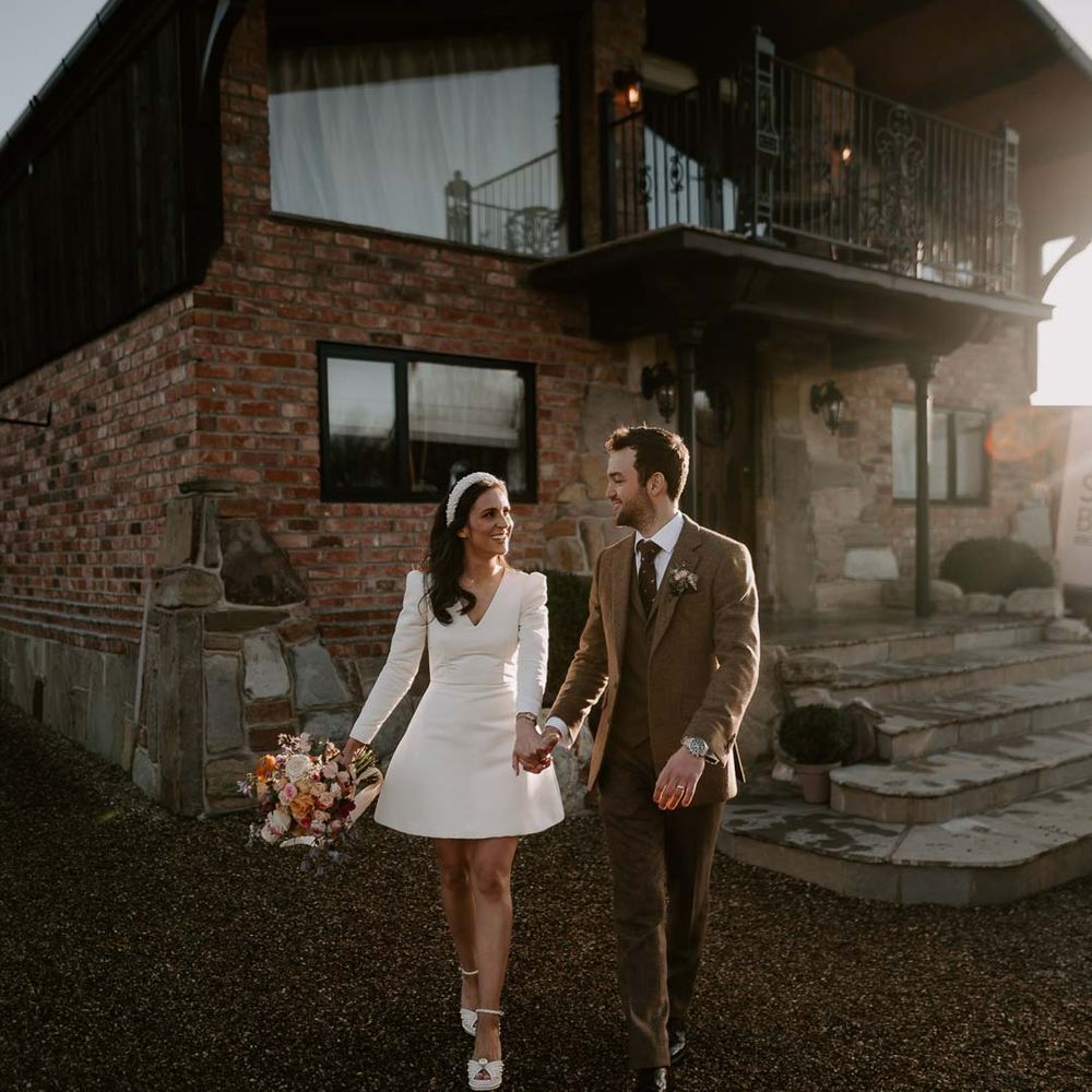 Groom in chocolate brown grooms suit walking with bride in long sleeve short wedding dress, pearl bridal headband and platform pearl wedding heels outside Willow Marsh Farm Loughborough wedding venue