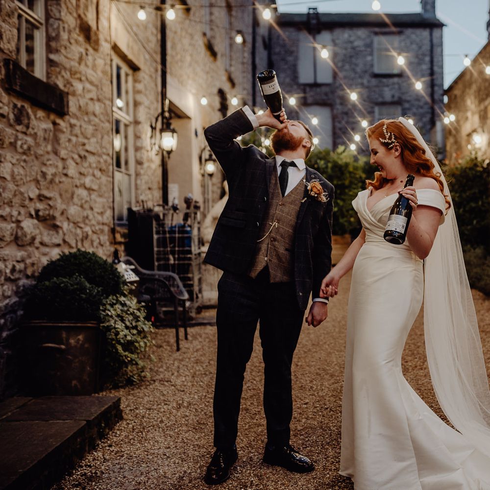 Bride and groom drinking champagne in Holmes Mill's Courtyard covered in festoon lights. 