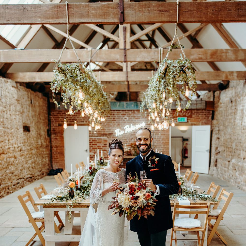 Bride & groom stand in rustic wedding venue with wooden tables and hanging lights