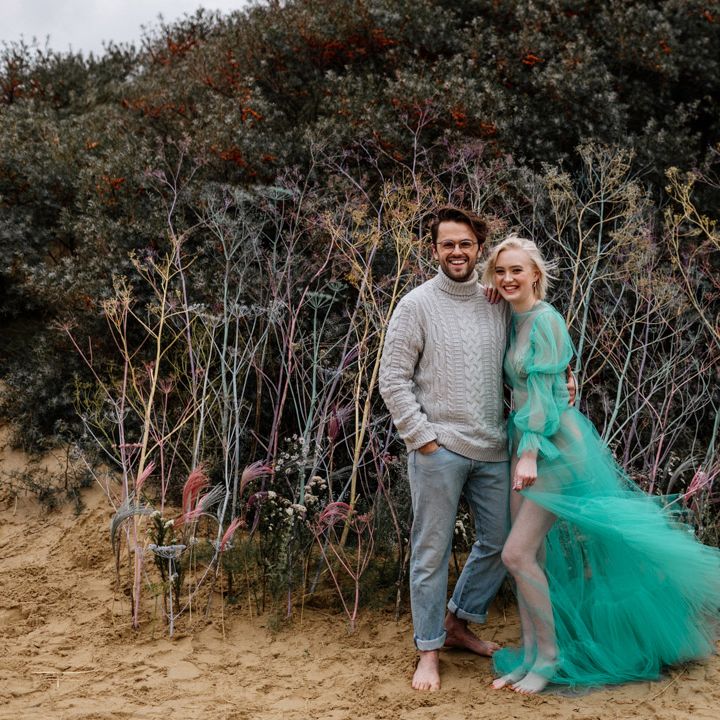 White bearded groom with glasses in a jumper and jeans on the beach with his bride in a sheer pastel wedding dress