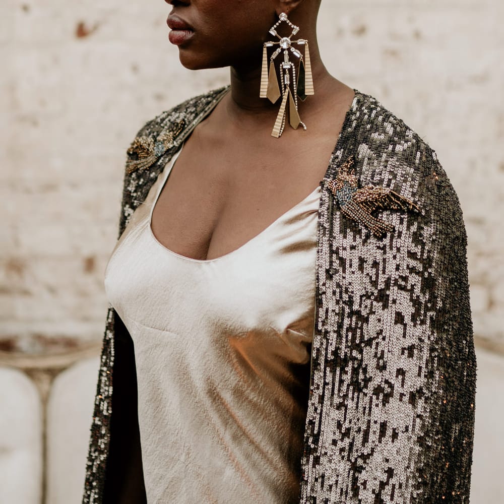 Stylish bride with alopecia in a satin wedding dress and black bridal cape with gold crown and statement earrings 