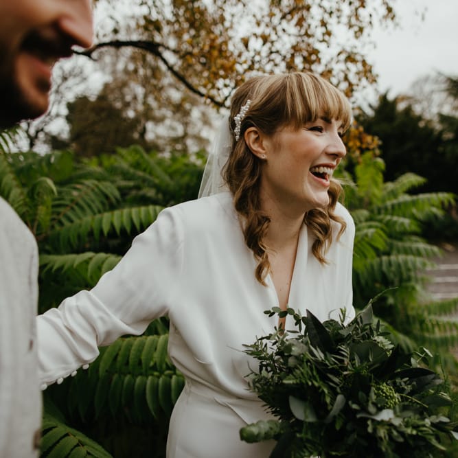 smiley bride in a wrap jumpsuit with blunt fringe and wavy hair holding a green foliage wedding bouquet