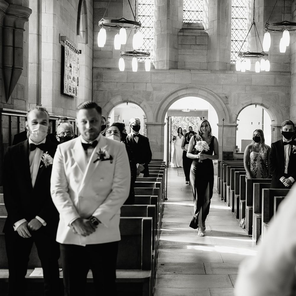 Bridesmaid in black dress holding white rose bouquet walks up the aisle towards groom in white double breasted tuxedo jacket and bow tie
