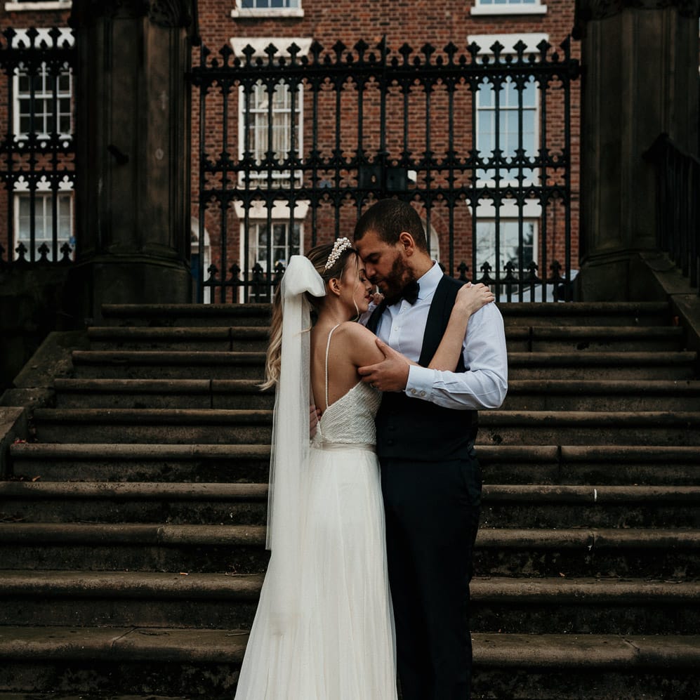 Groom in a horseshoe waistcoat and bow tie embracing his bride in a thin strap wedding dress with bow veil 