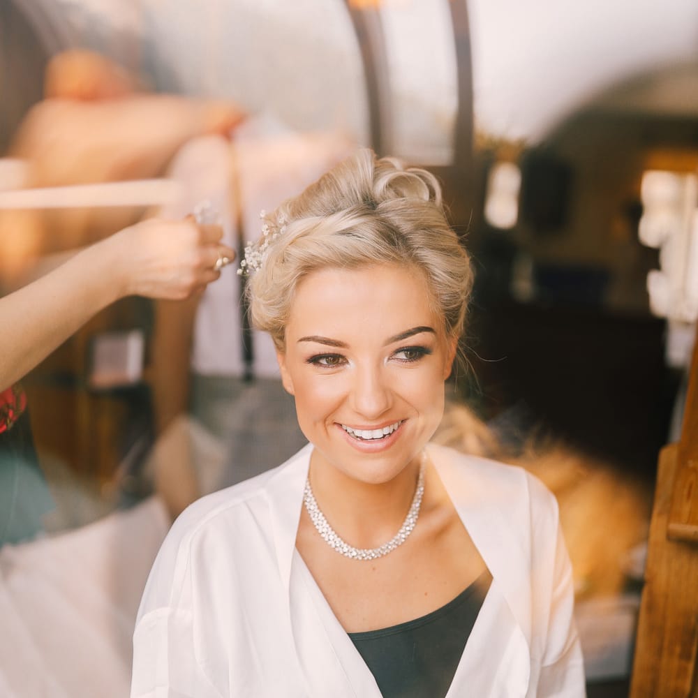 A blonde bride wearing a white robe gets gypsophelia put into her hair as she gets ready for her wedding. 