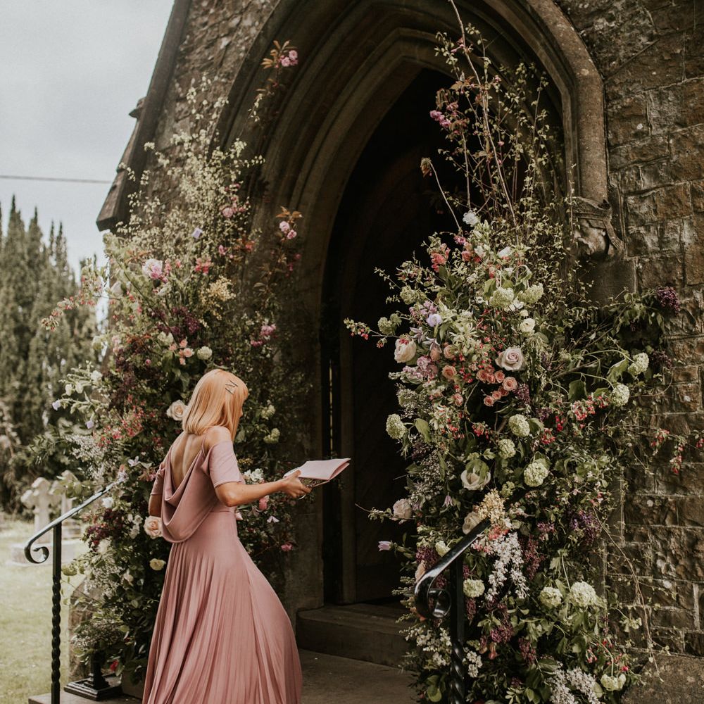 Bridesmaid in a pink cowl back dress walking into the church decorated with wild flower arrangements
