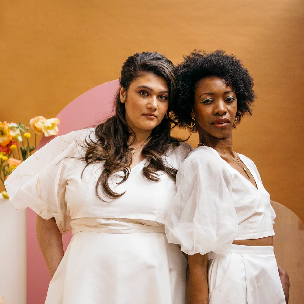 Two brides leaning on each other wearing matching Paper To Lace wedding separates with puff sleeves and long maxi skirt