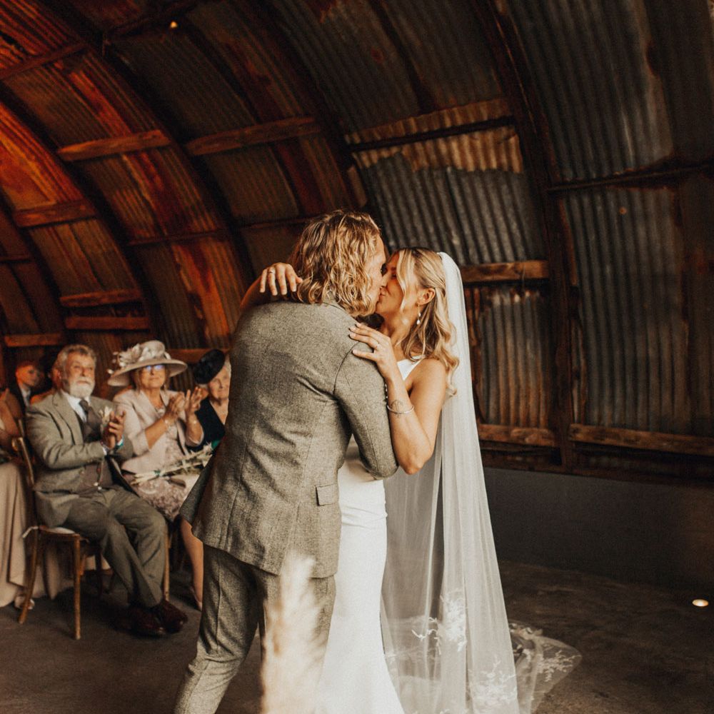 Bride in white Made With Love wedding dress and embroidered veil kisses groom in sage green suit and brown shoes during wedding ceremony at Anran Devon