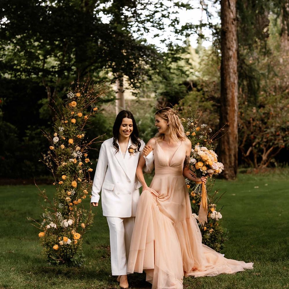 Two brides in a white trouser suit and blush wedding dress walking up the outdoor aisle with yellow vertical flower arrangement 