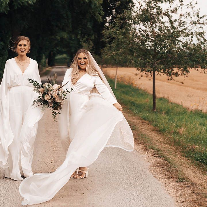Brides walk along together as bride wears cape with jumpsuit and blonde bride wears veil