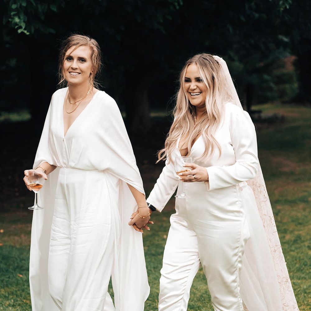 Brides walk along grass in bridal jumpsuits on their wedding day