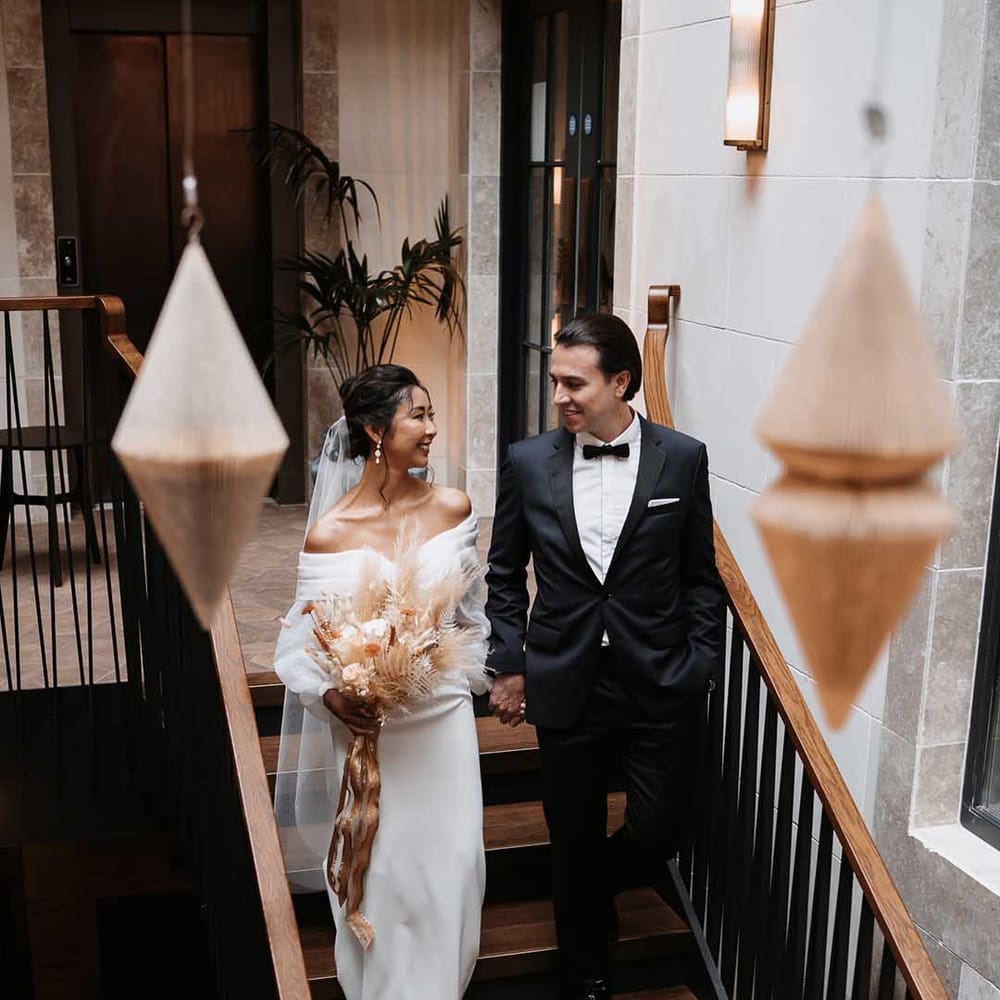 Bride holding dried blush pink tone and nude flower arrangement holding groom in tailored fitted black suit 
