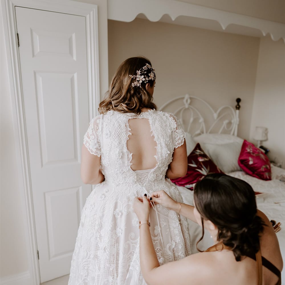 Bride in homemade lace wedding dress with open back has the back fastened by bridesmaid in rust cross back satin bridesmaid dress before DIY garden wedding in Bedfordshire
