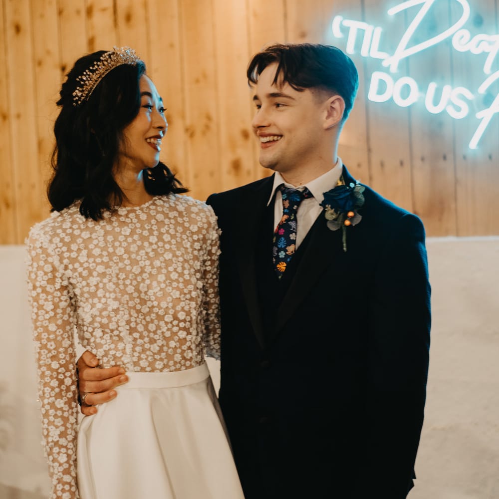 Bride in long sleeve applique wedding dress and headpiece stands with groom in dark blue suit, floral tie and floral buttonhole stand by neon sign at rustic wedding breakfast 