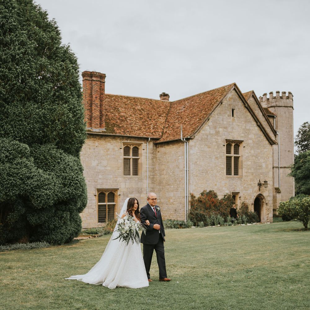 Bride in Pronovias wedding dress, Jimmy Choo wedding shoes and veil carrying white rose and green bridal bouquet walks arm in arm with man in dark suit and white rose buttonhole towards the aisle at outdoor wedding ceremony