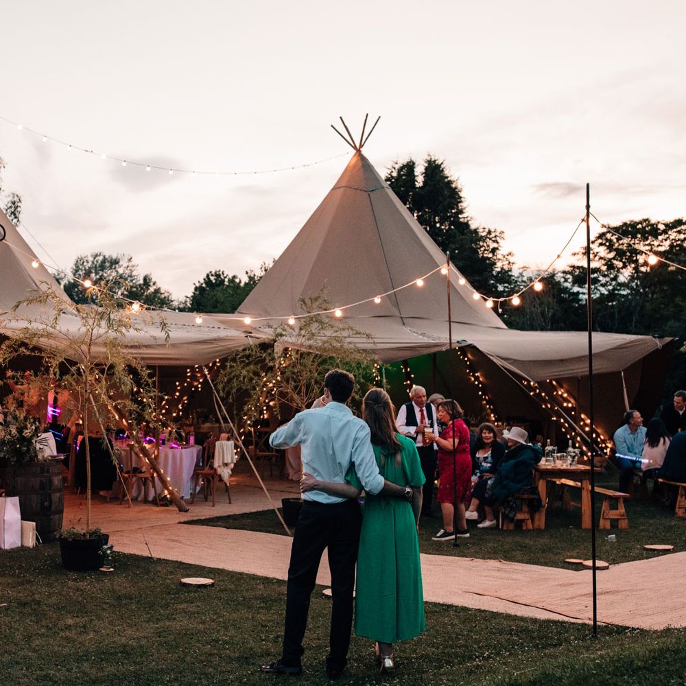 Wedding guests watch on as guests dance in fairy lit tipi for festival style wedding reception during golden hour