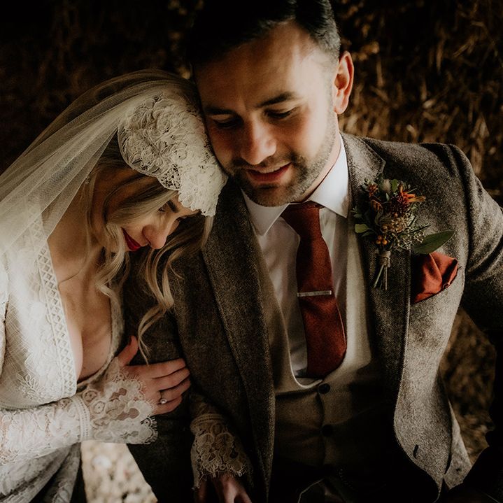 Bride in a lace wedding dress and Juliet cap veil leaning on her husbands shoulder in a brown wool suit