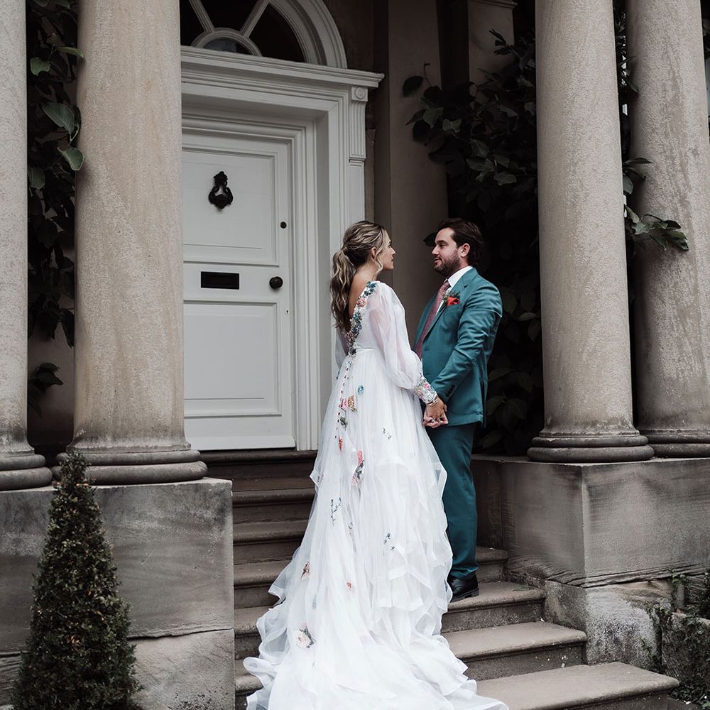 Bride in low back long sleeve embroidered wedding dress with train stands on stone steps holding hands with groom in teal suit
