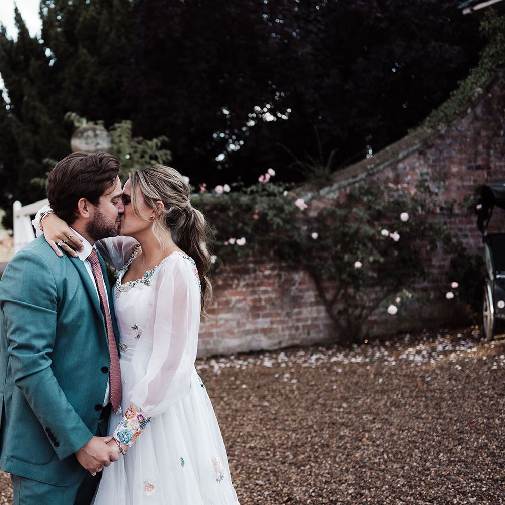 Groom in blue suit and pink tie kisses bride in long sleeve Victoria Sanders wedding dress with embroidered cuffs in the grounds of Iscoyd Park