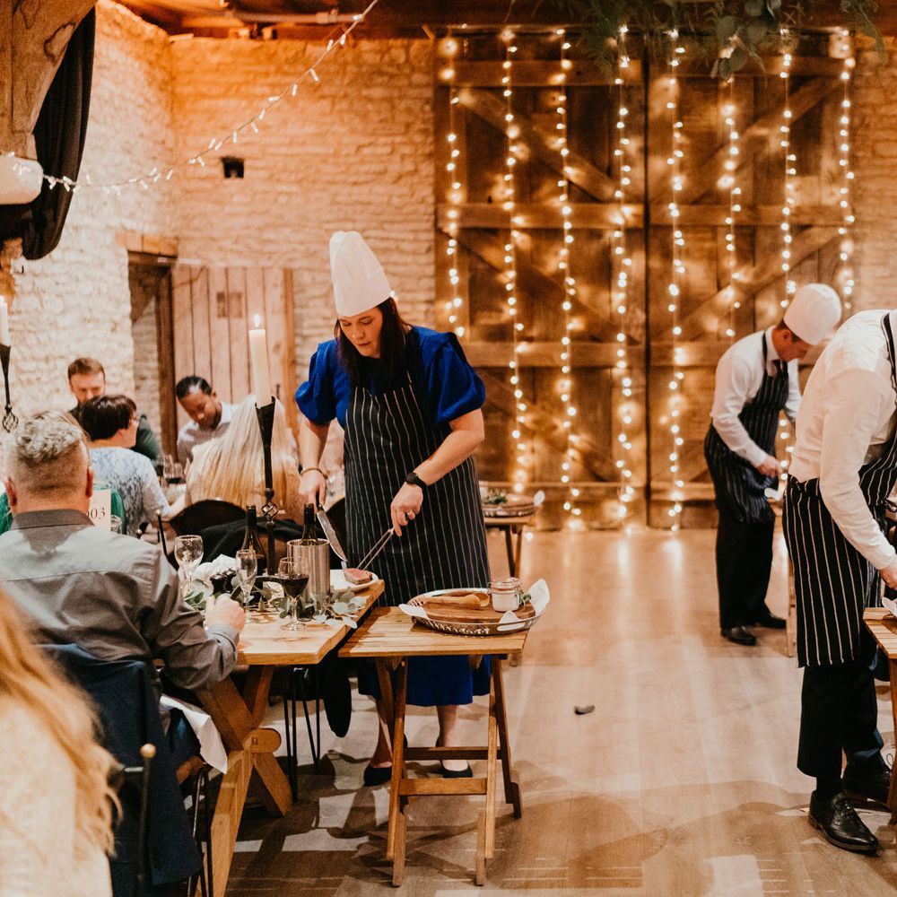 Guests in striped aprons and chefs hats cut up meats at tables for barn wedding reception with fairy light canopy