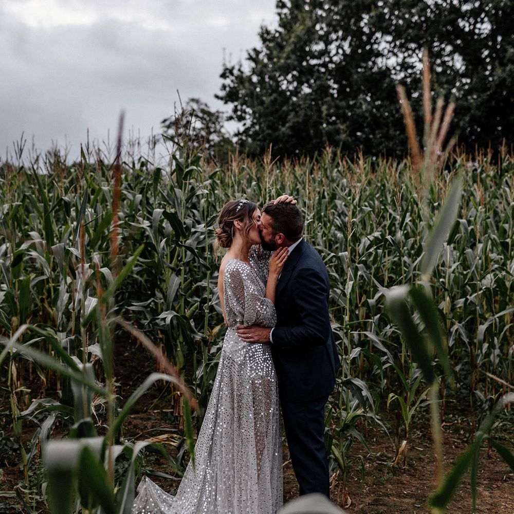Bride in a silver sequin wedding dress and groom in a navy suit kissing amongst the green crops 