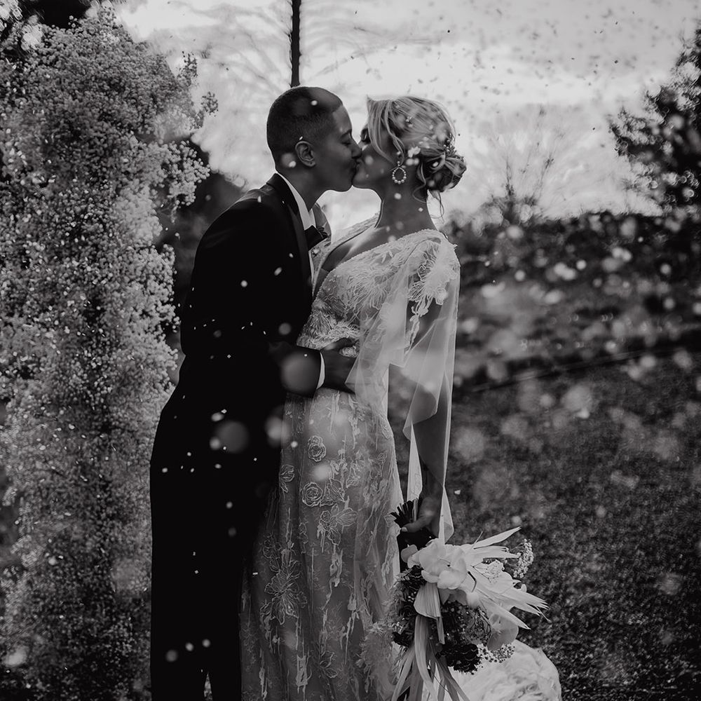 Black and white confetti portrait of an LGBTQI+ couple kissing in a black tuxedo and lace wedding dress at Hodsock Priory