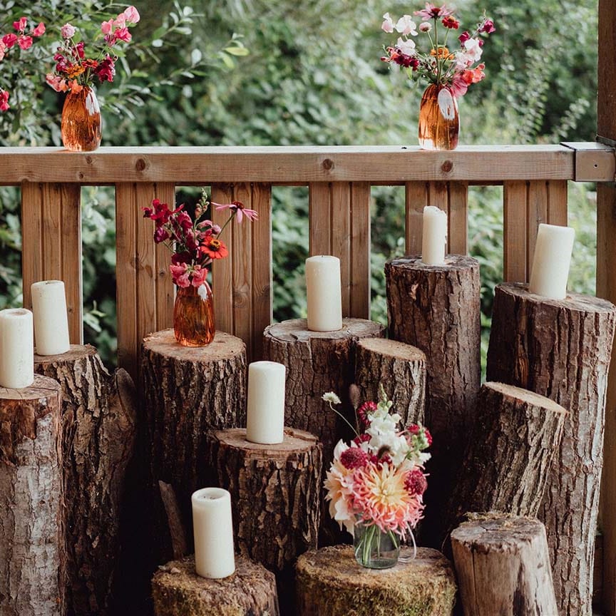 Wedding decor of different sized tree trunks with white pillar candles and colourful flowers in orange glass vases