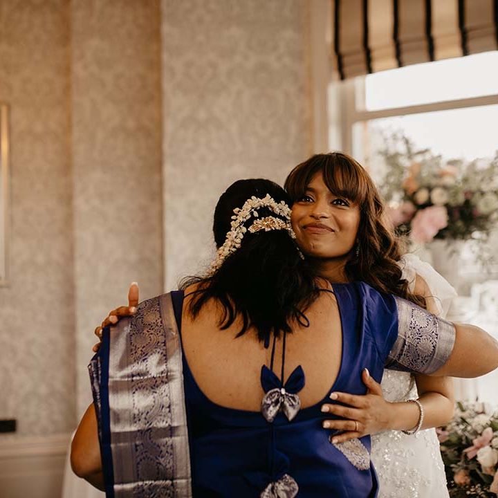 Bride in white traditional wedding dress embracing wedding guest in deep purple dress with gold embellishments