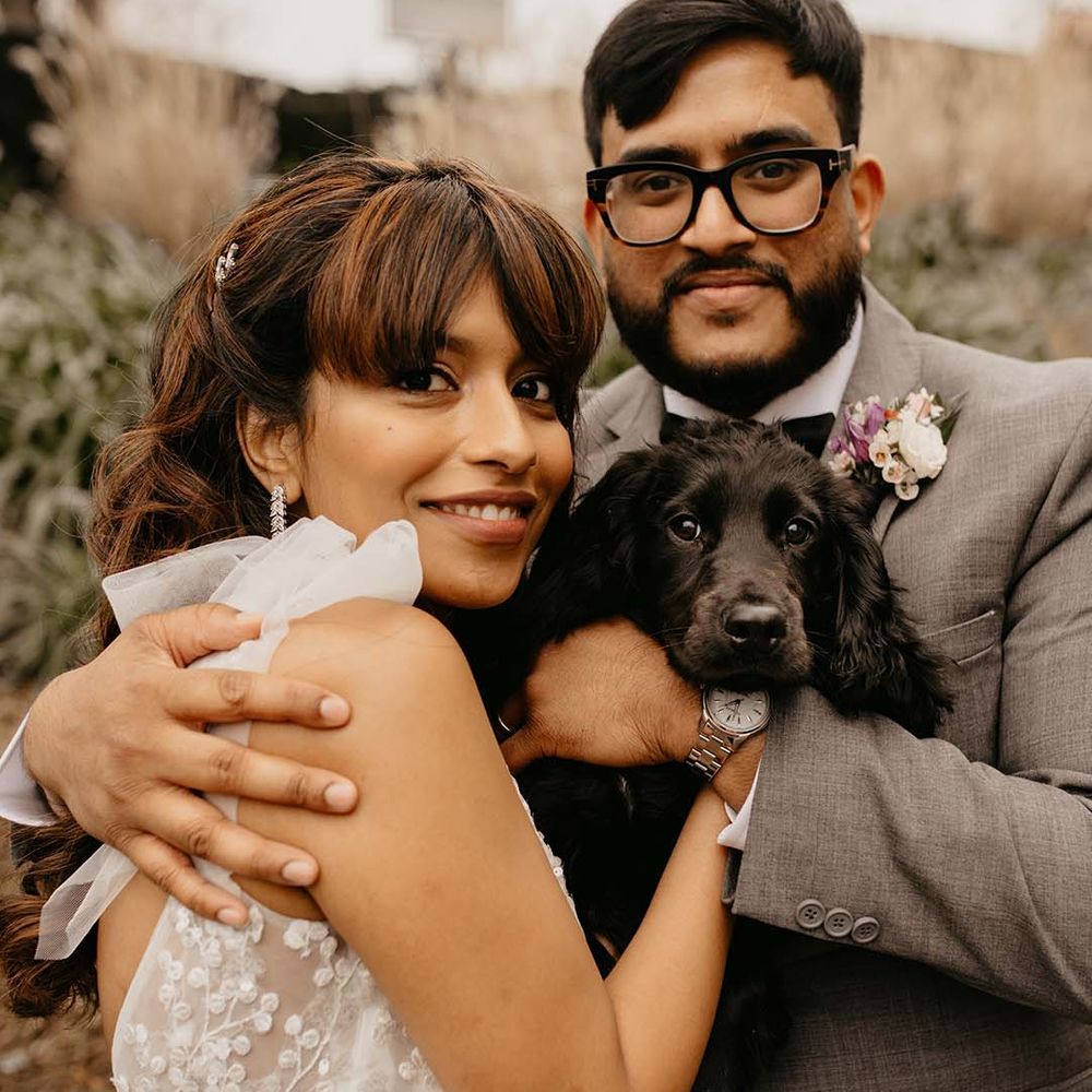 Bride in white traditional wedding dress with silver dangling earrings holding black puppy with groom in grey suit with bow tie and boutonniere 