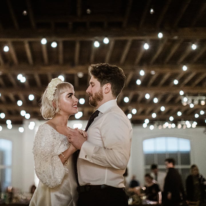 Bride with short blonde hair in a short wedding dress and white flower headband has first dance with groom in shirt and suit trousers