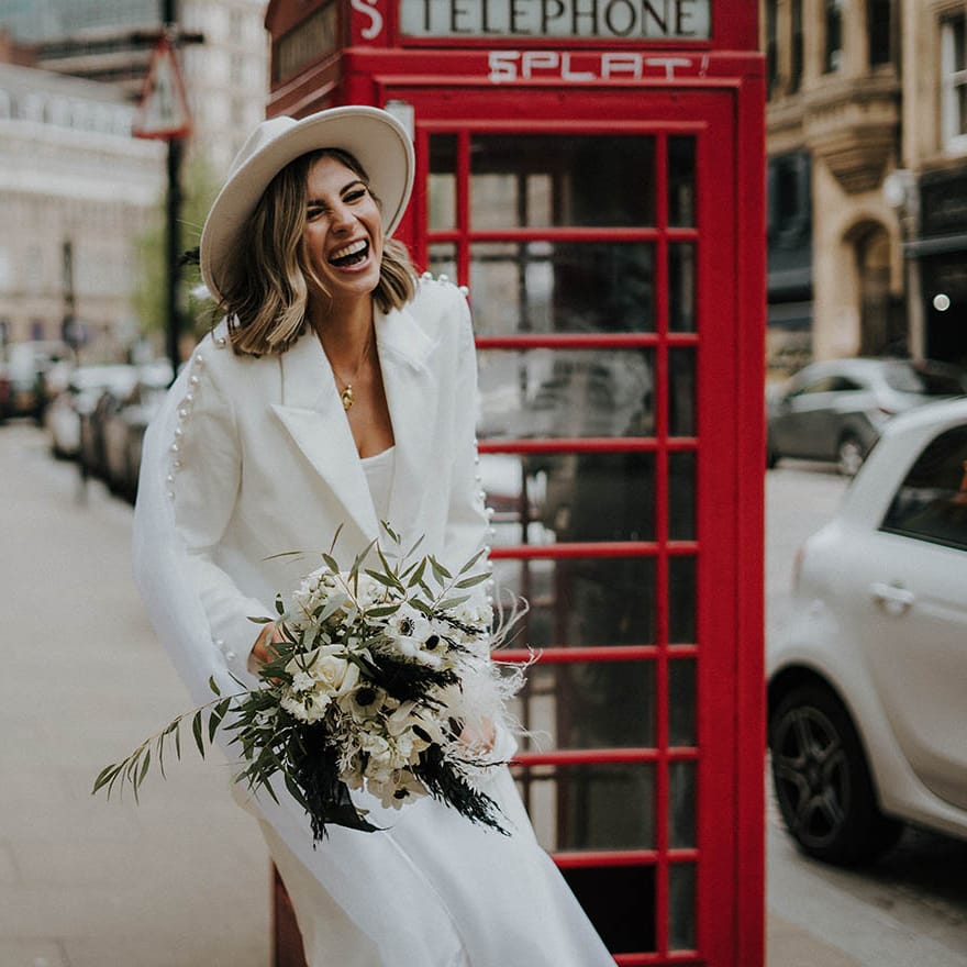 Bride laughing wearing a fedora and wedding suit with pearls with white flower bouquet