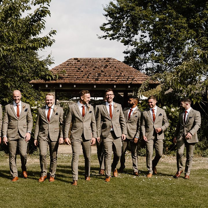 Groom walks with groomsmen in matching suits and orange ties for multicultural wedding at the Great Tythe Barn Tetbury