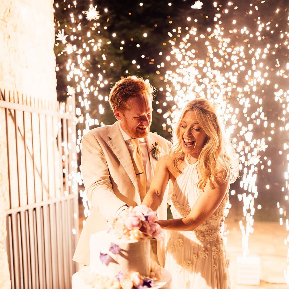 Bride and groom cut their three tier rustic wedding cake decorated with purple flowers as fireworks explode
