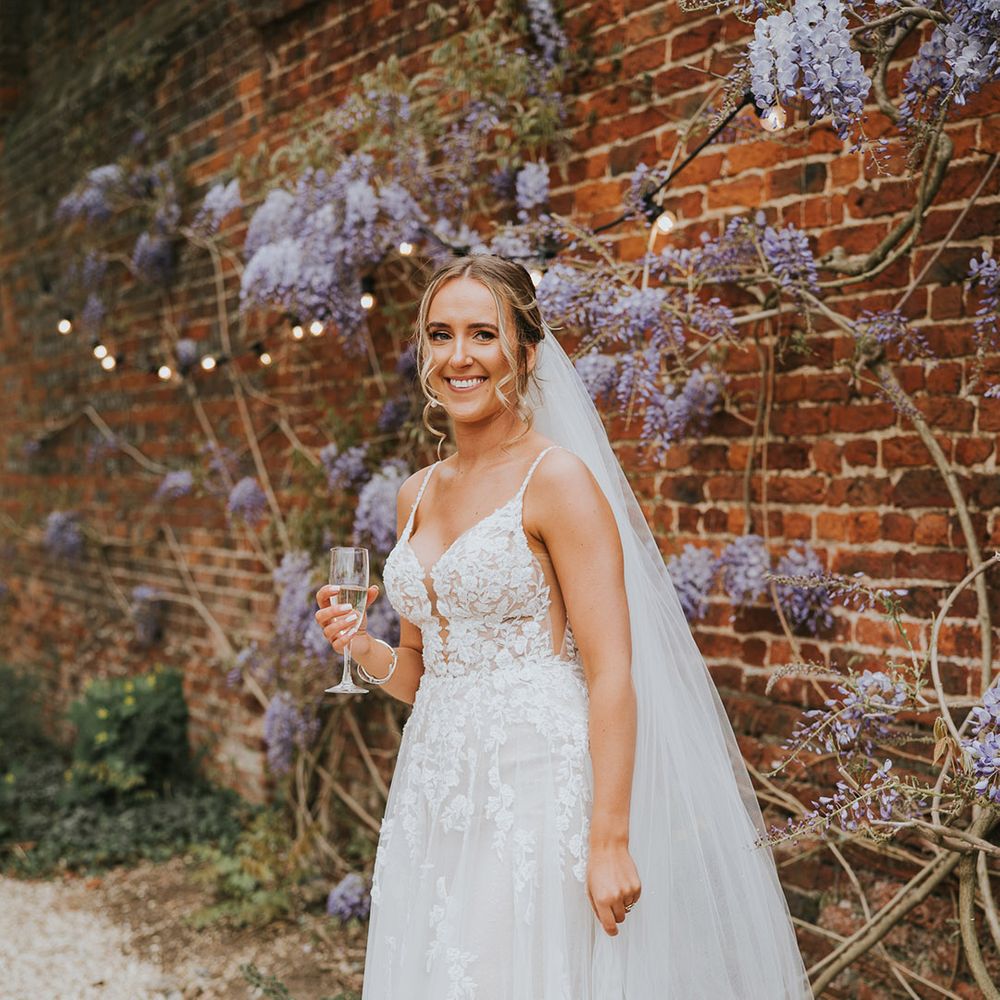 Lace wedding dress with thin straps holding a glass of champagne 